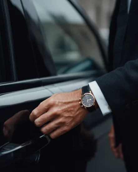 A hand wearing a dress watch resting on the open door of a black car